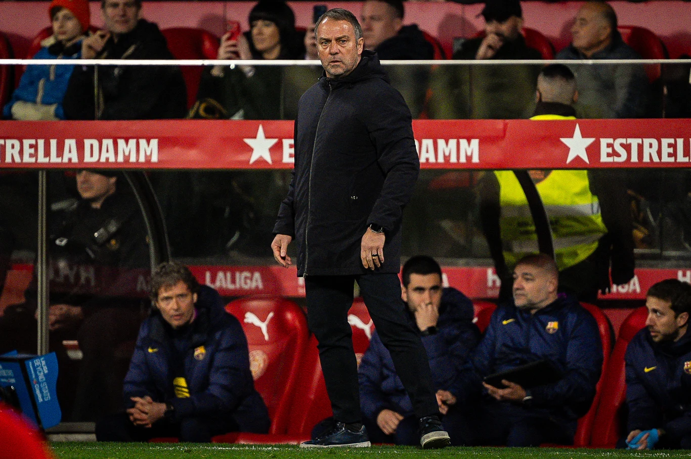 Trener w ciemnej kurtce stoi przy linii bocznej boiska piłkarskiego, za nim ławka rezerwowych i kilku członków sztabu szkoleniowego, na trybunach widoczni kibice i czerwone bannery z napisem Estrella Damm.