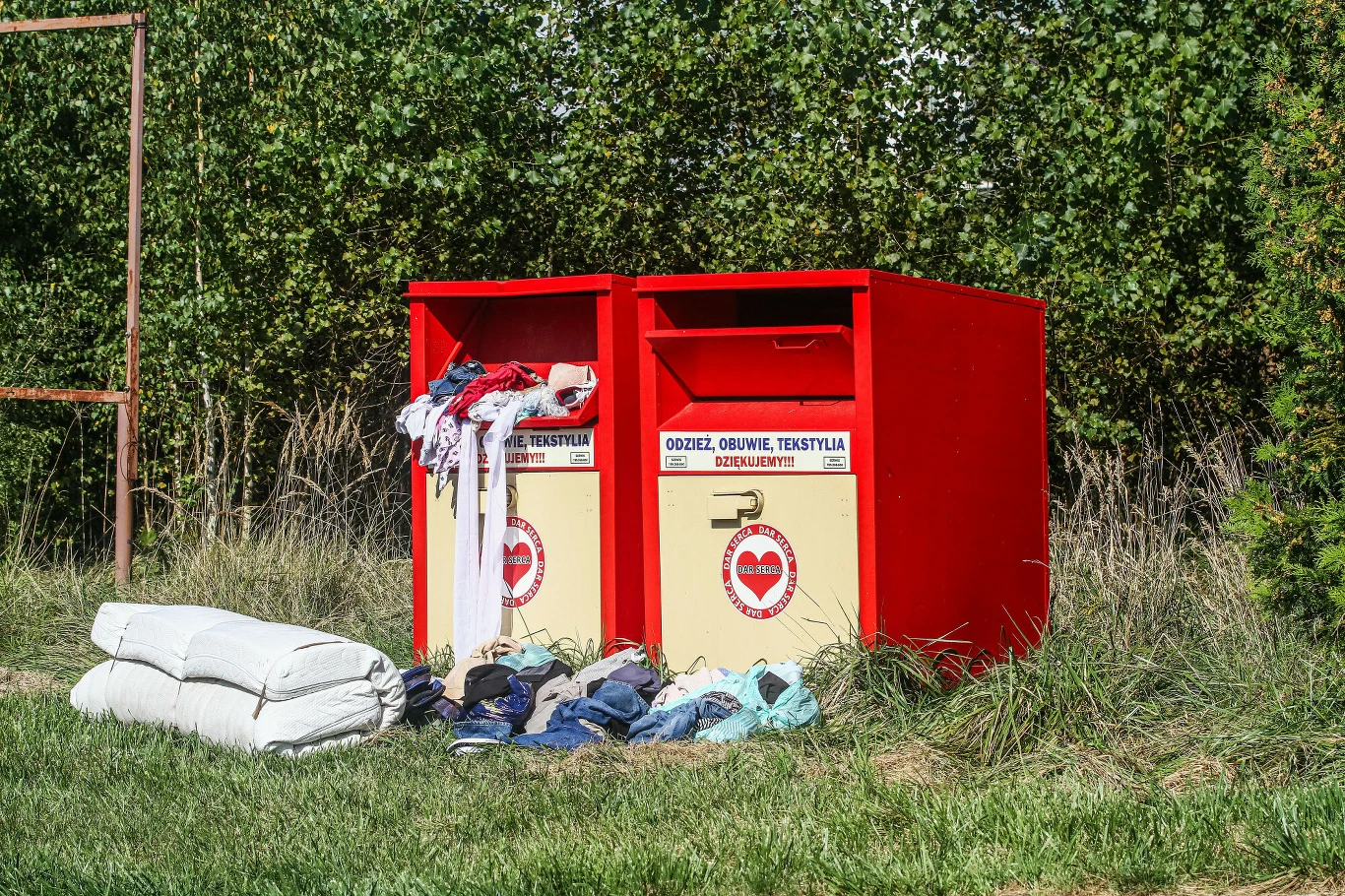 Od kwietnia 2028 r. producenci tekstyliów w ramach ROP mają zacząć płacić za ich późniejsze zagospodarowanie, gdy staną się odpadami Dwa czerwone pojemniki oznaczone do zbiórki odzieży używanej, na których widoczne jest logo organizacji charytatywnej. Przed pojemnikami leżą porozrzucane ubrania, tekstylia i poduszki, otoczone zieloną trawą oraz drzewami.