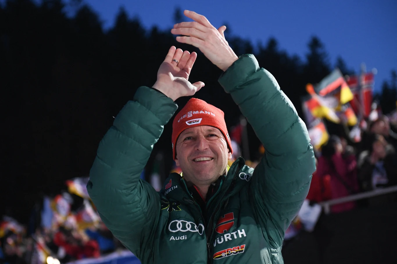 Mężczyzna w zielonej kurtce oraz czerwonej czapce z napisem, uśmiecha się i unosi ręce do góry w geście aplauzu, w tle widoczne są flagi oraz rozmyta publiczność na tle ciemnych drzew.