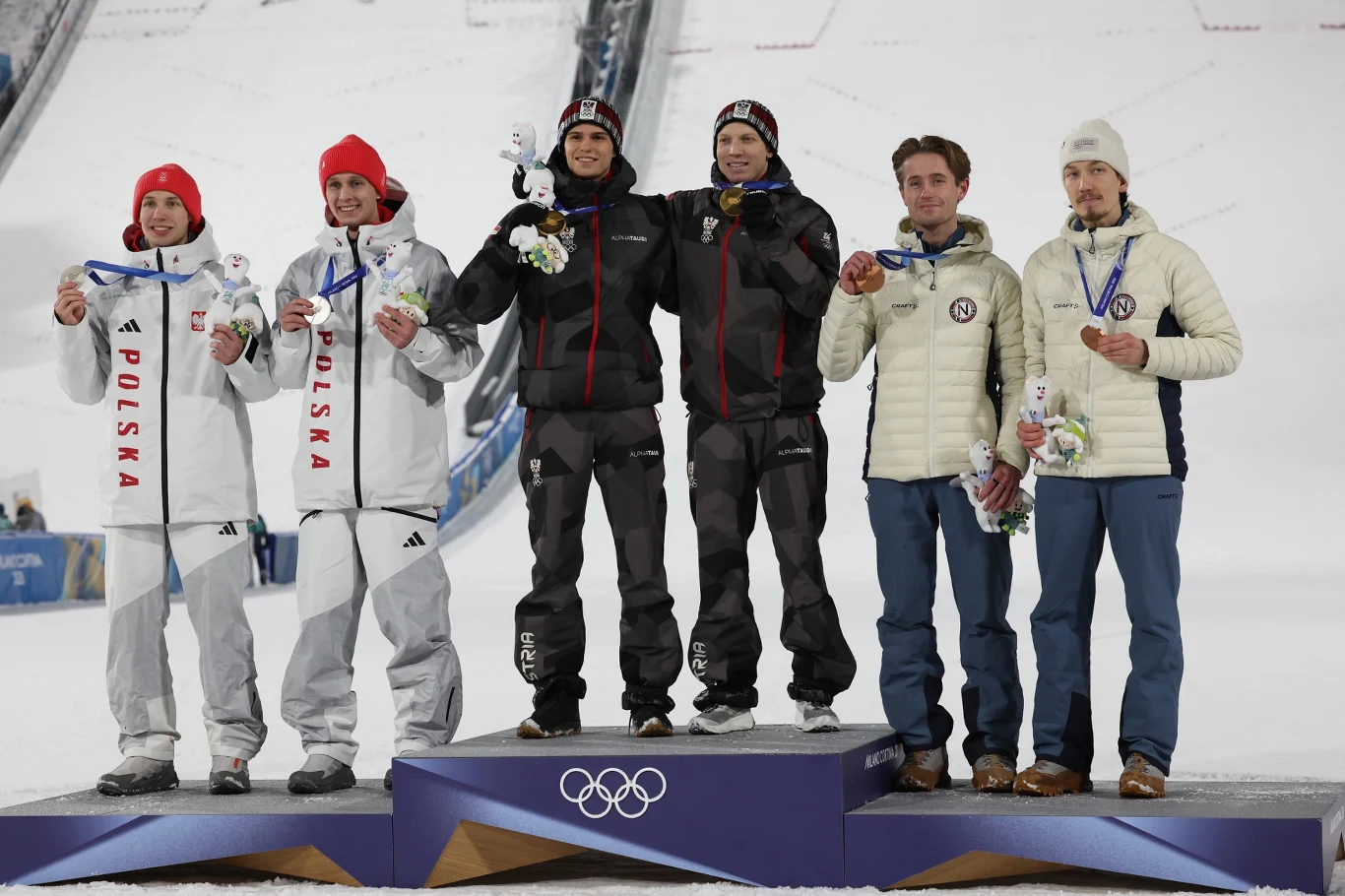 Trzy drużyny sportowców stoją na podium na tle śnieżnej skoczni narciarskiej, trzymając medale i kwiaty. Zawodnicy ubrani są w oficjalne stroje reprezentacji olimpijskich, dumnie prezentują swoje osiągnięcia.