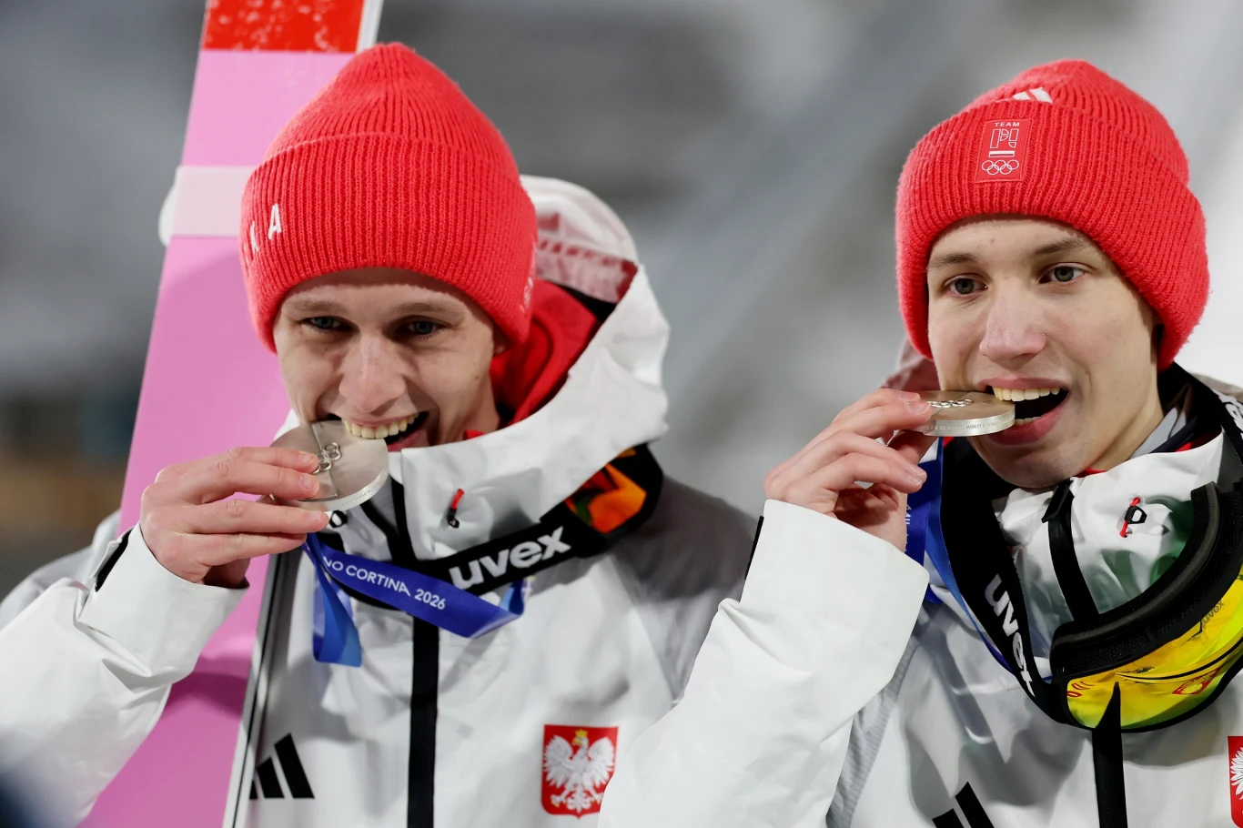 Dwóch sportowców w białych kurtkach z polskim godłem i czerwonych czapkach smakuje medale. Mają na sobie akredytacje i cieszą się z sukcesu na tle zimowej scenerii.