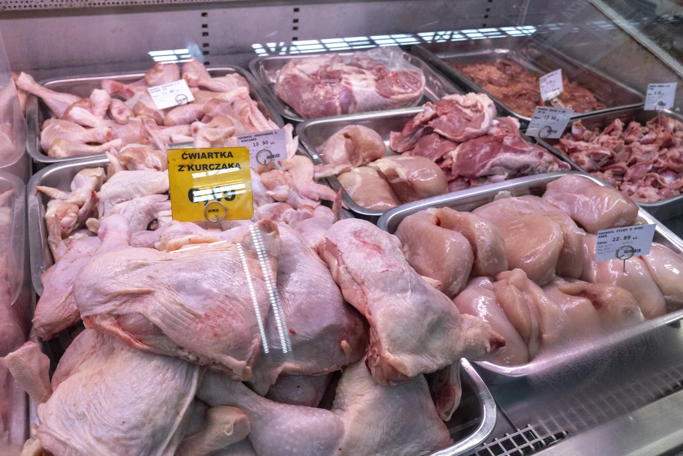 Różne rodzaje surowego mięsa drobiowego ułożone w metalowych pojemnikach za szybą chłodni w sklepie, z widoczną żółtą etykietą informującą o ćwiartce z kurczaka.