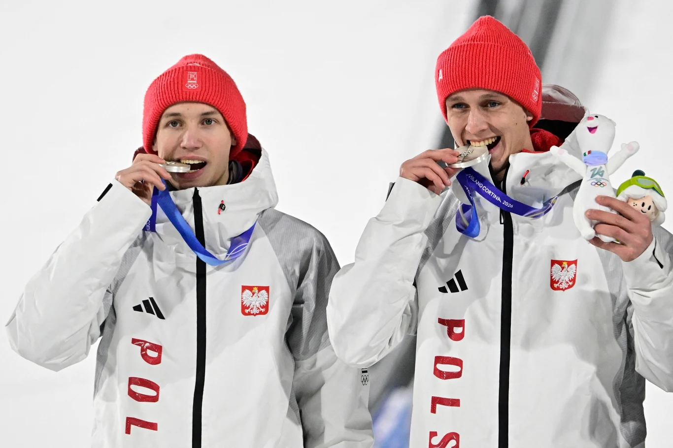 Dwóch sportowców w białych kurtkach z polskimi godłami i czerwonych czapkach gryzie medale, mają na szyi niebieskie wstążki oraz uśmiechnięte twarze. Jeden z nich trzyma maskotkę.