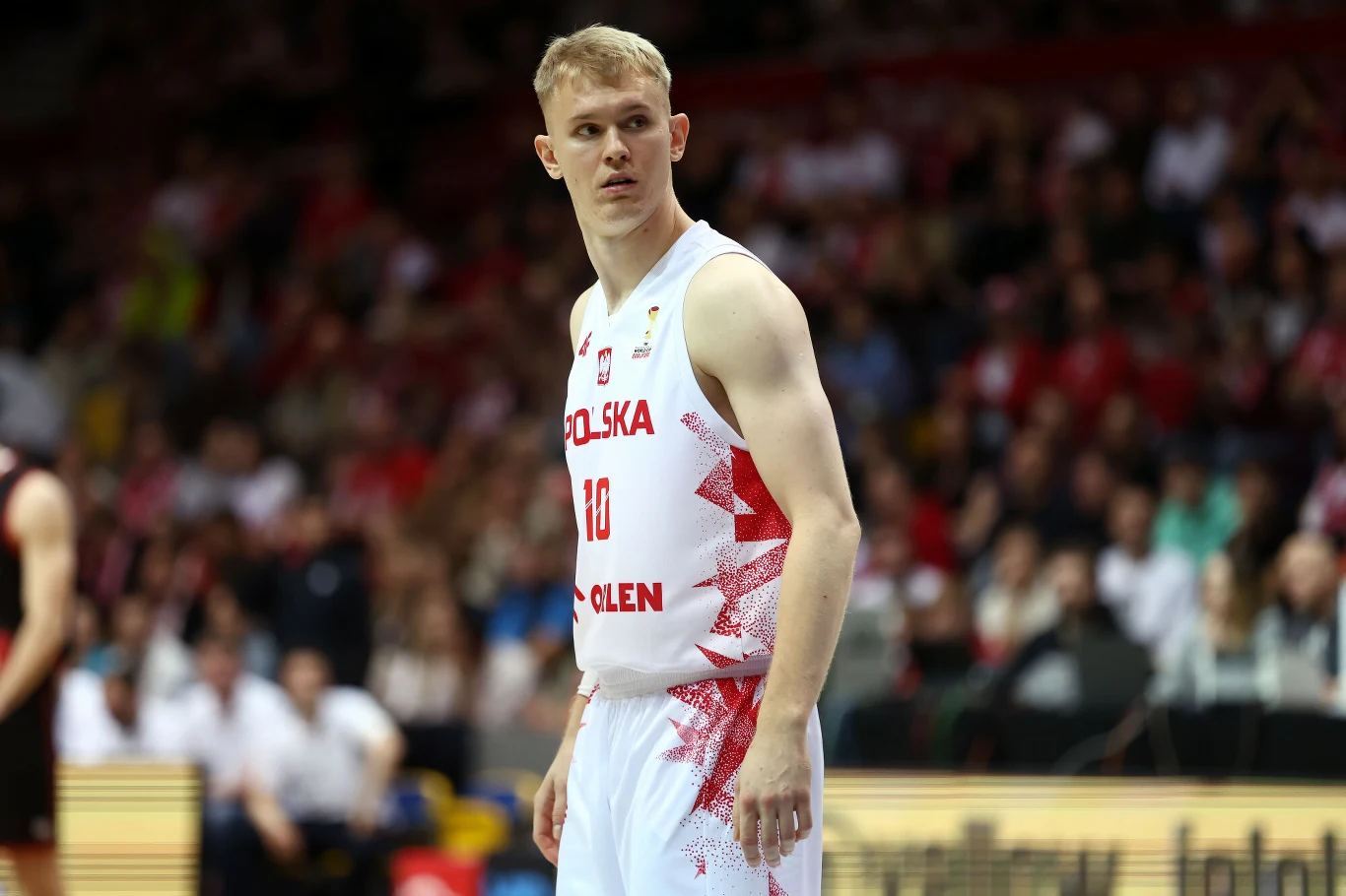 Młody koszykarz ubrany w biały strój z napisem Polska i numerem 10 stoi na boisku podczas meczu, w tle widoczni kibice na trybunach.