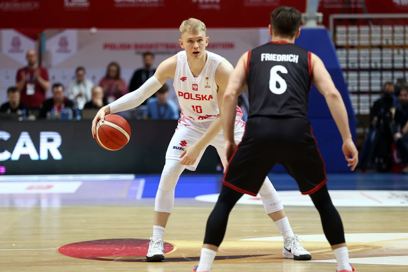 Dwóch zawodników drużyn narodowych Polski i Niemiec rywalizuje podczas meczu koszykówki na hali sportowej, jeden z nich kozłuje piłkę, drugi broni.