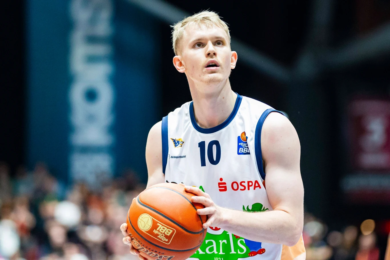 Koszykarz przygotowuje się do wykonania rzutu wolnego podczas meczu, skupiony na celu. Ubrany w białą koszulkę z numerem 10 oraz logotypami sponsorów, trzyma piłkę do koszykówki. W tle widoczni rozmyci kibice oraz fragment hali sportowej.