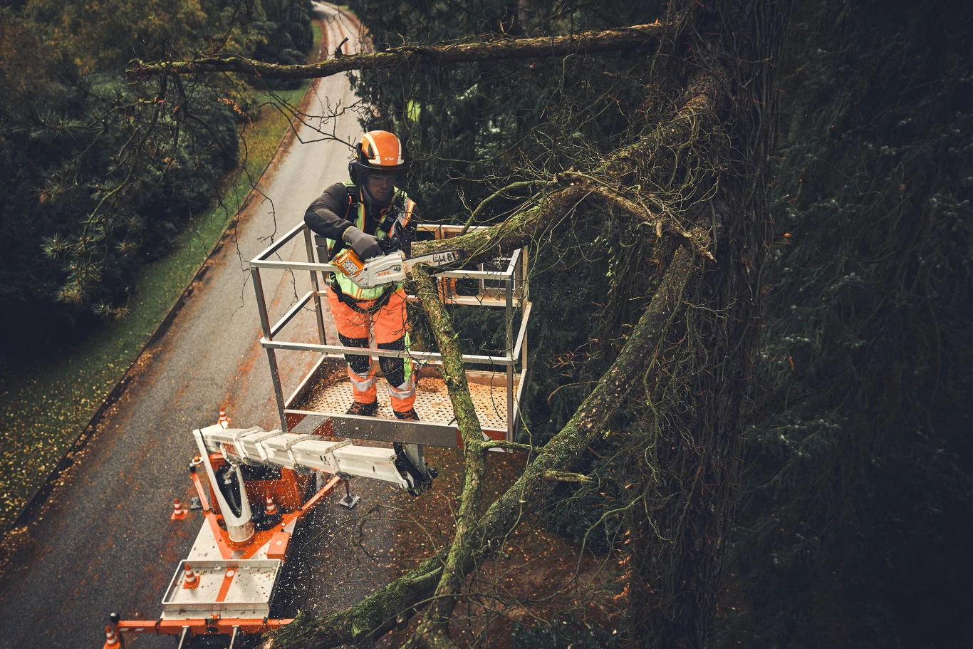 Pracownik w odzieży ochronnej przycina gałęzie drzewa z podnośnika koszowego przy ruchliwej drodze, używając piły mechanicznej. Gałęzie są duże i rozłożyste. Widoczny profesjonalny sprzęt i środki bezpieczeństwa.