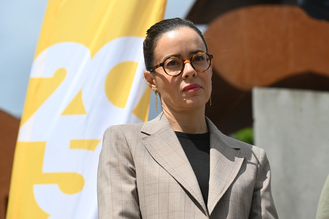 Aleksandra Leo rozważa odejście z Polski 2050. "Trudno sobie wyobrazić" Kobieta w okularach i marynarce stoi przed żółtym banerem z widocznymi białymi cyframi. Tło stanowią rozmyte elementy architektury i fragmenty niebieskiego nieba.