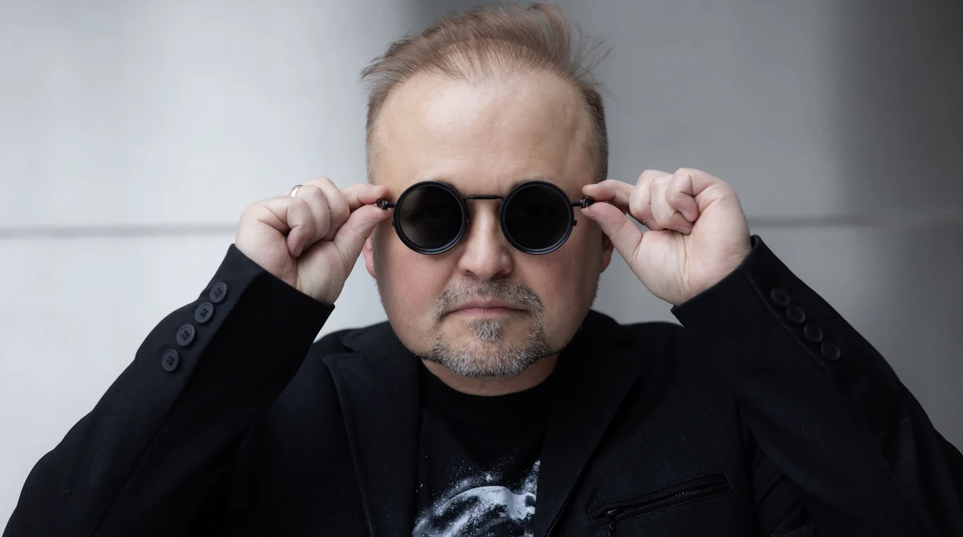 Maciej Zieliński Mężczyzna w okrągłych, czarnych okularach przeciwsłonecznych, ubrany w ciemną marynarkę i czarną koszulkę z nadrukiem, trzyma okulary oburącz i patrzy prosto w obiektyw. Jego fryzura jest częściowo przerzedzona, tło jasnoszare i neutralne.