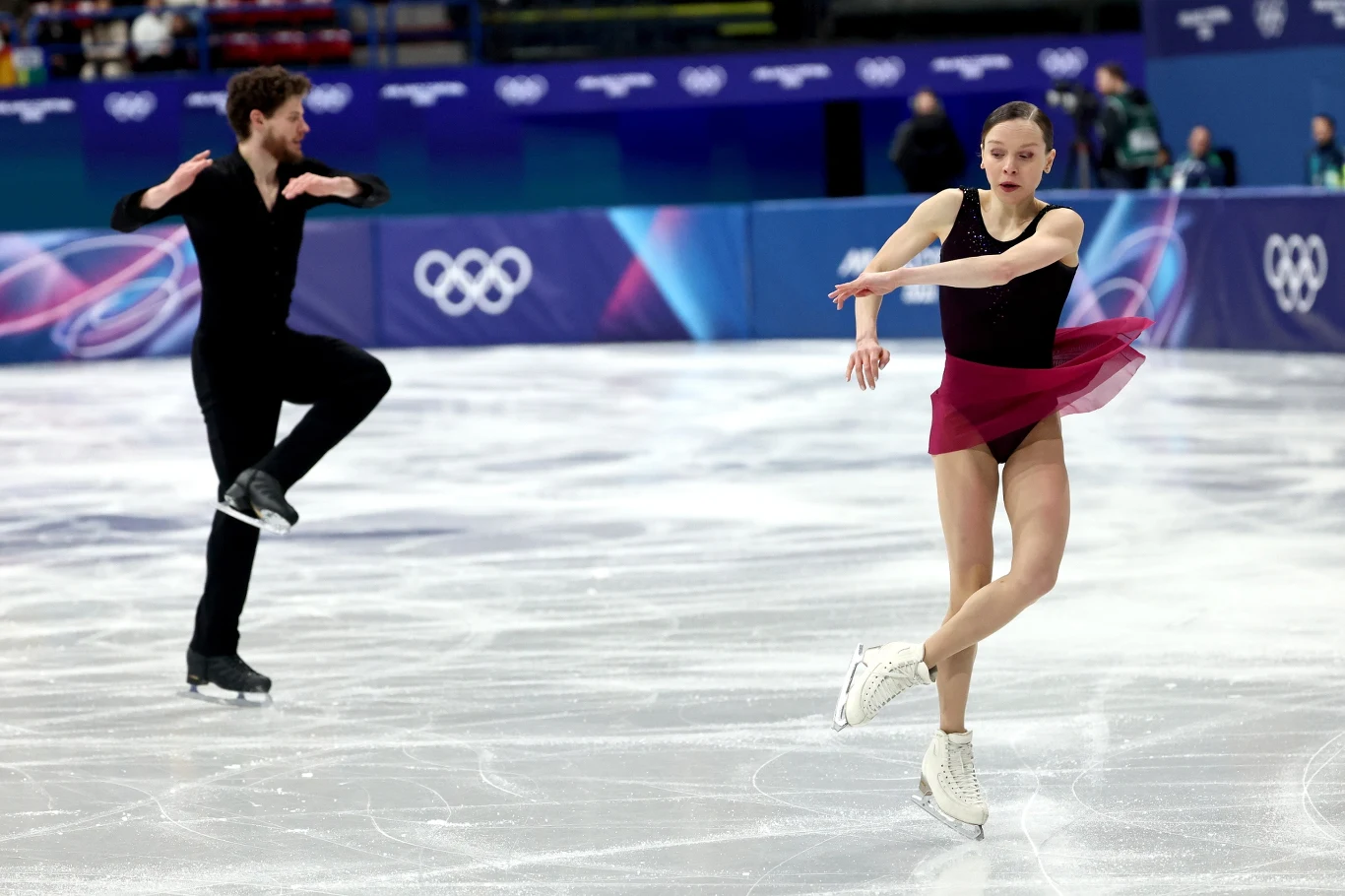 Dwoje łyżwiarzy figurowych wykonuje program na lodowisku podczas zawodów olimpijskich. Kobieta w bordowej spódniczce i czarnej bluzce unosi rękę, mężczyzna w czarnym stroju w tle jest w ruchu. W tle widoczne są logotypy olimpijskie i widownia.