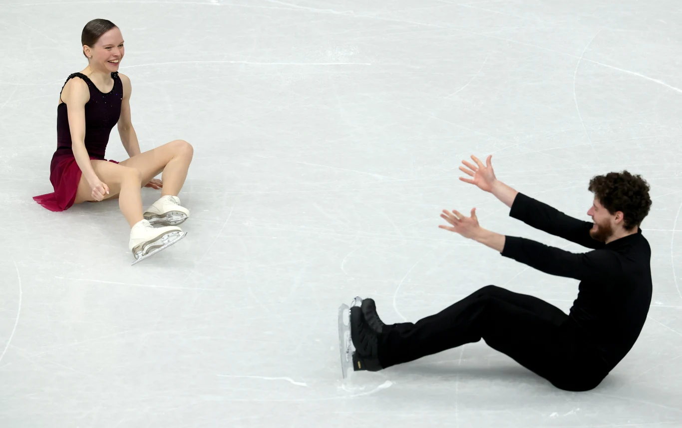 Dwoje łyżwiarzy figurowych siedzi na lodowisku naprzeciwko siebie, uśmiechając się do siebie nawzajem po upadku podczas występu.