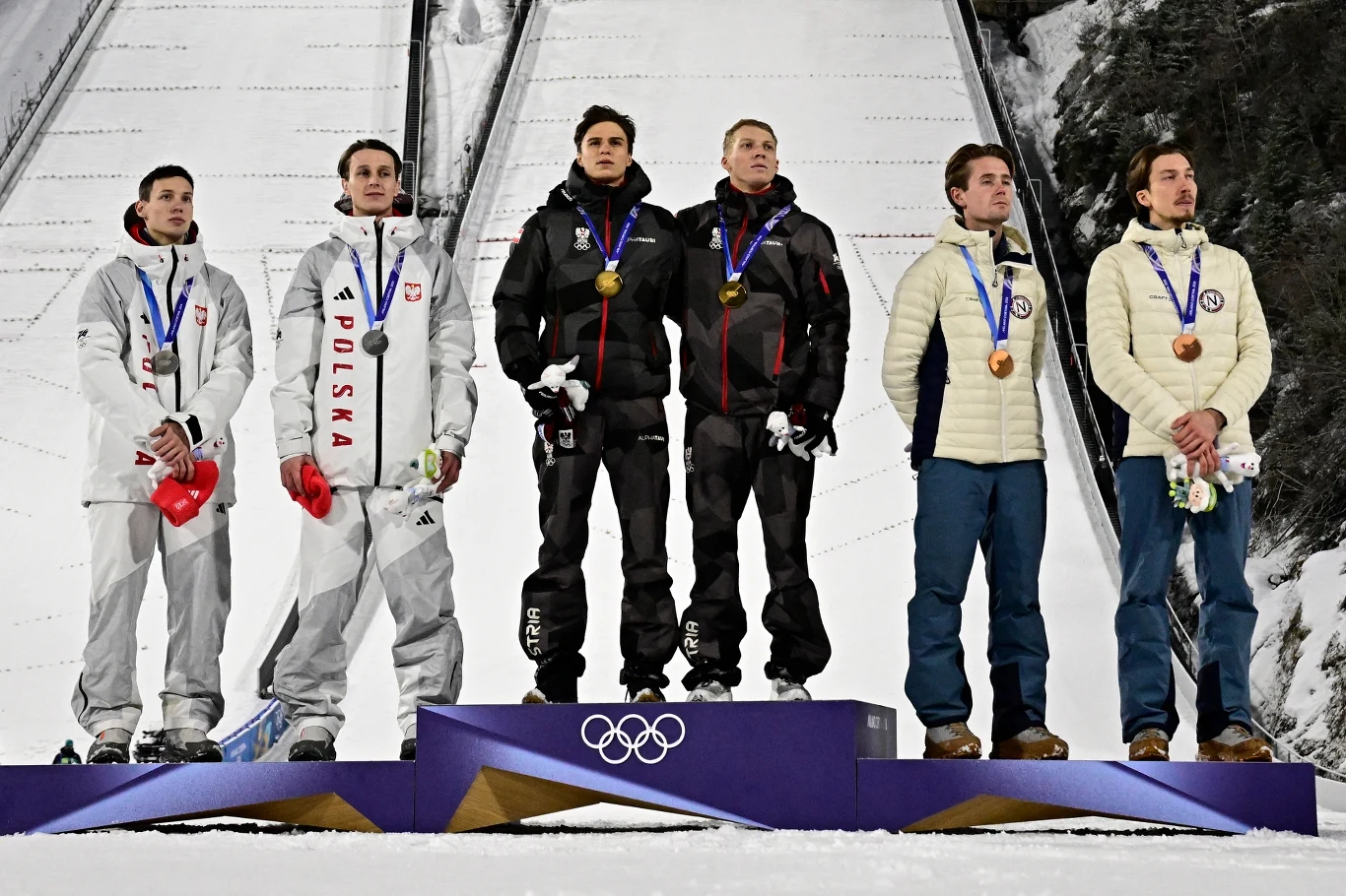 Sześciu młodych mężczyzn stojących na podium olimpijskim, ubrani w stroje narodowe z medalami na szyjach, tło stanowi skocznia narciarska i zimowy krajobraz.