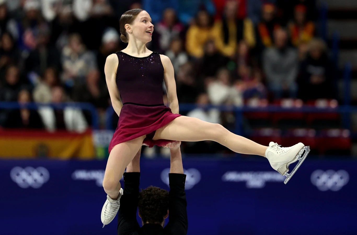 Łyżwiarka figurowa w bordowej sukience podczas podnoszenia przez partnera na lodowisku. W tle widoczni są kibice oraz nieostre flagi i logo igrzysk olimpijskich.