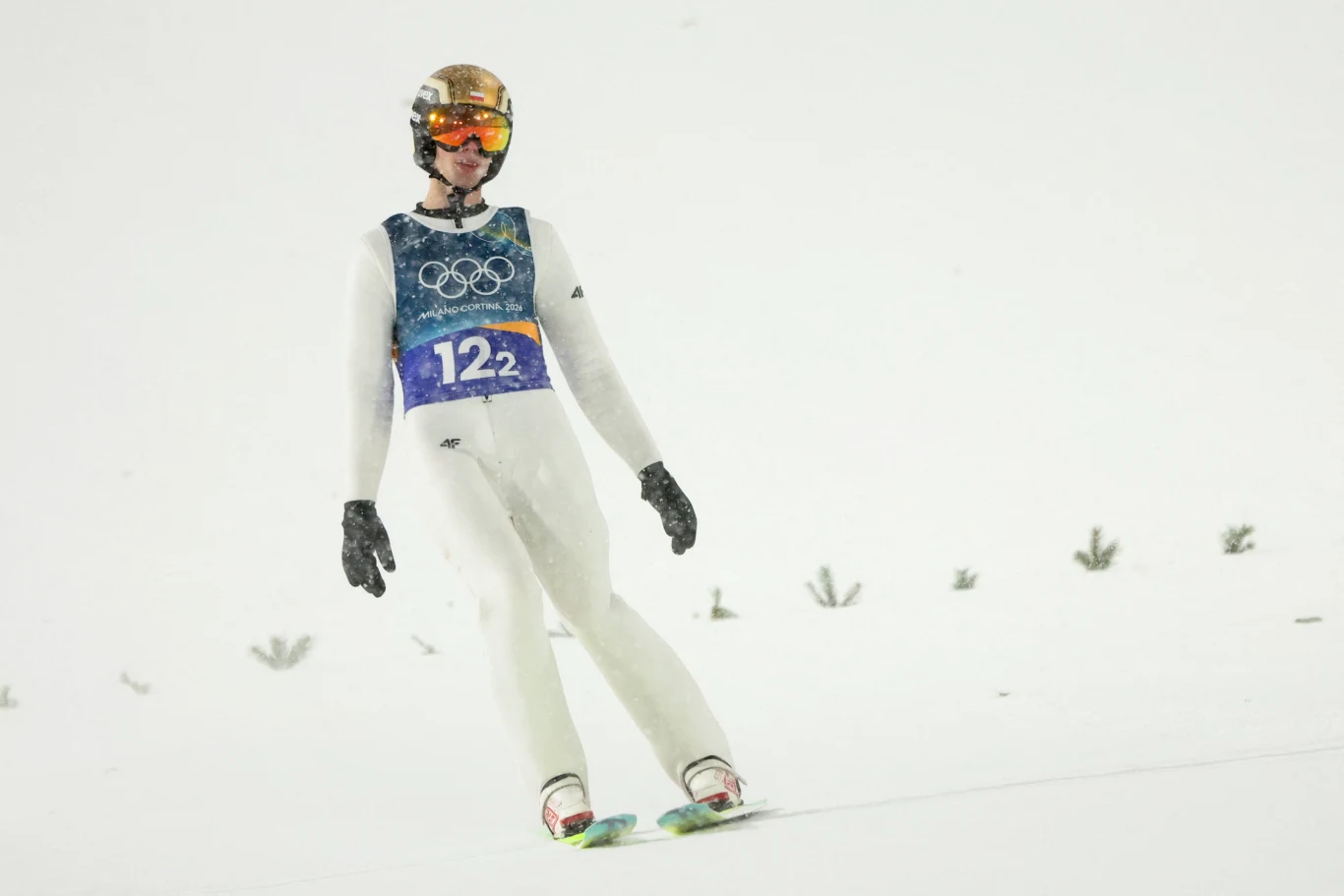 Skoczek narciarski w stroju sportowym ląduje na śnieżnej skoczni podczas zawodów olimpijskich, widoczne są elementy wyposażenia oraz numer startowy 122.