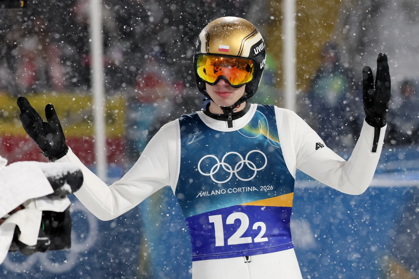 Kacper Tomasiak Skoczek narciarski w kombinezonie i kasku z goglami podczas zimowych igrzysk olimpijskich w śnieżnej scenerii, z uniesionymi rękami.