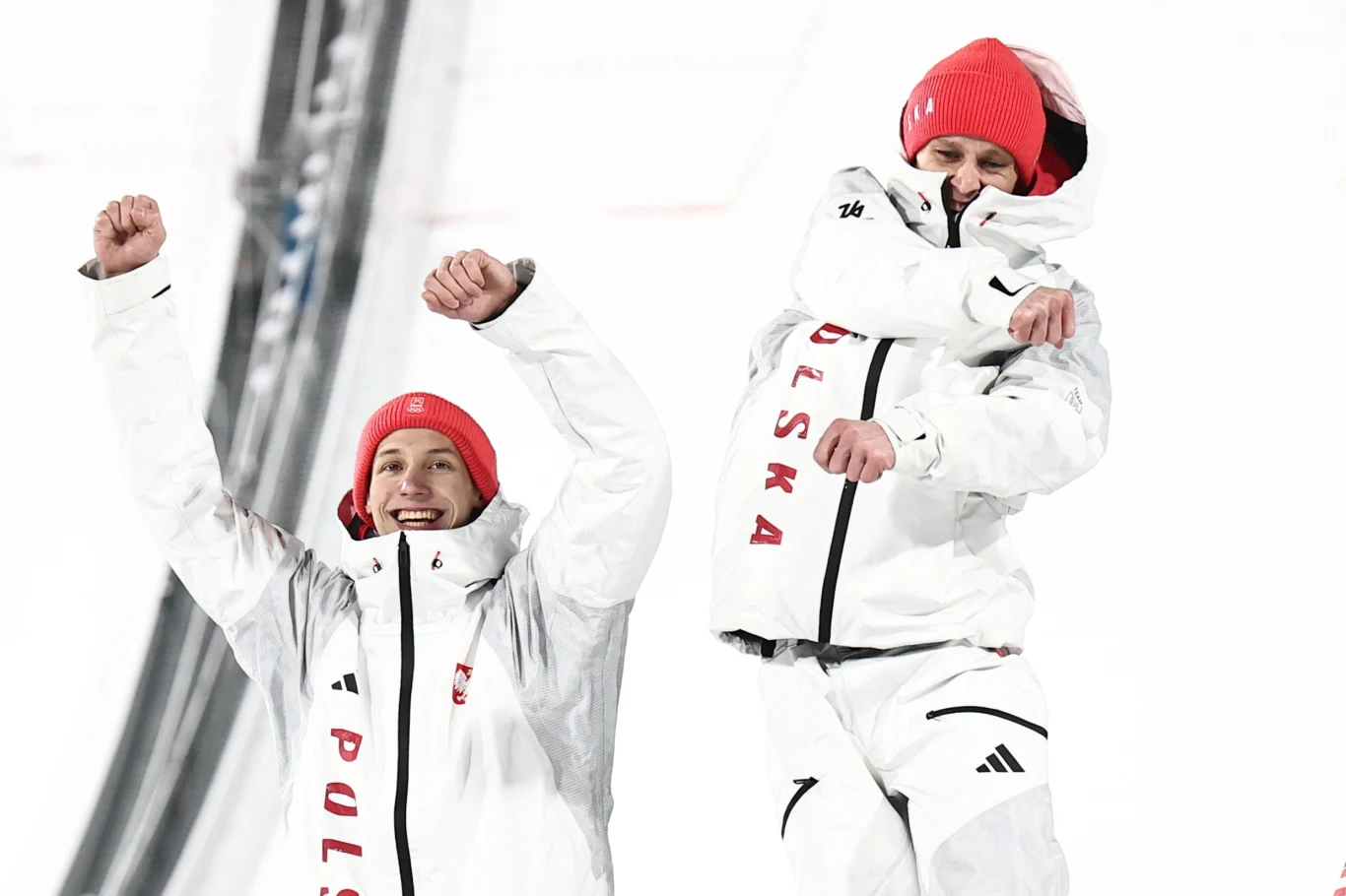 Kacper Tomasiak i Paweł Wąsek Dwóch sportowców w białych kurtkach z czerwonym napisem 'Polska' oraz czerwonych czapkach cieszy się z sukcesu na tle śnieżnego obiektu sportowego.
