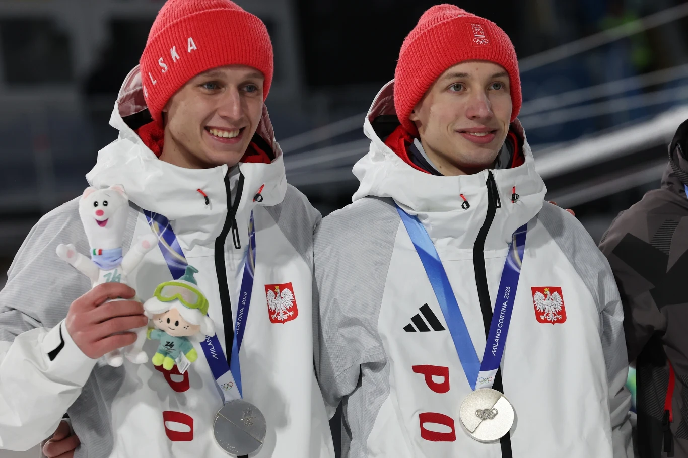 Kacper Tomasiak i Paweł Wąsek (po lewej) Dwóch młodych sportowców ubranych w białe kurtki z napisem Polska i czerwonych czapkach zimowych, stojących obok siebie. Każdy z nich ma na szyi medal olimpijski, a jeden trzyma maskotkę i pluszową zabawkę.