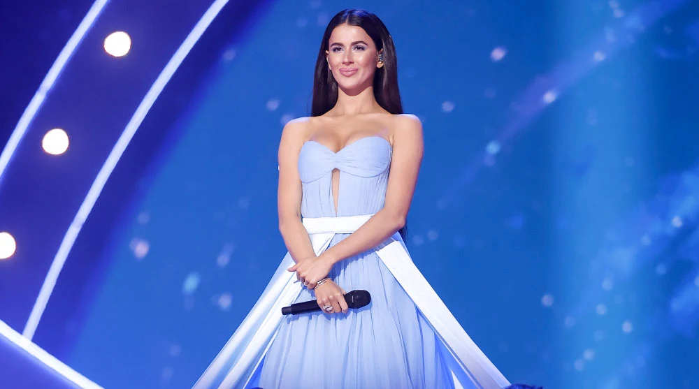 Roxie Węgiel nakręciła teledysk na zamarzniętym Bałtyku Młoda kobieta w długiej, jasnoniebieskiej sukni stoi na scenie na tle efektownego, rozświetlonego błękitem tła. Ma długie, ciemne włosy i lekko uśmiechnięty wyraz twarzy, a w dłoniach trzyma czarny mikrofon.