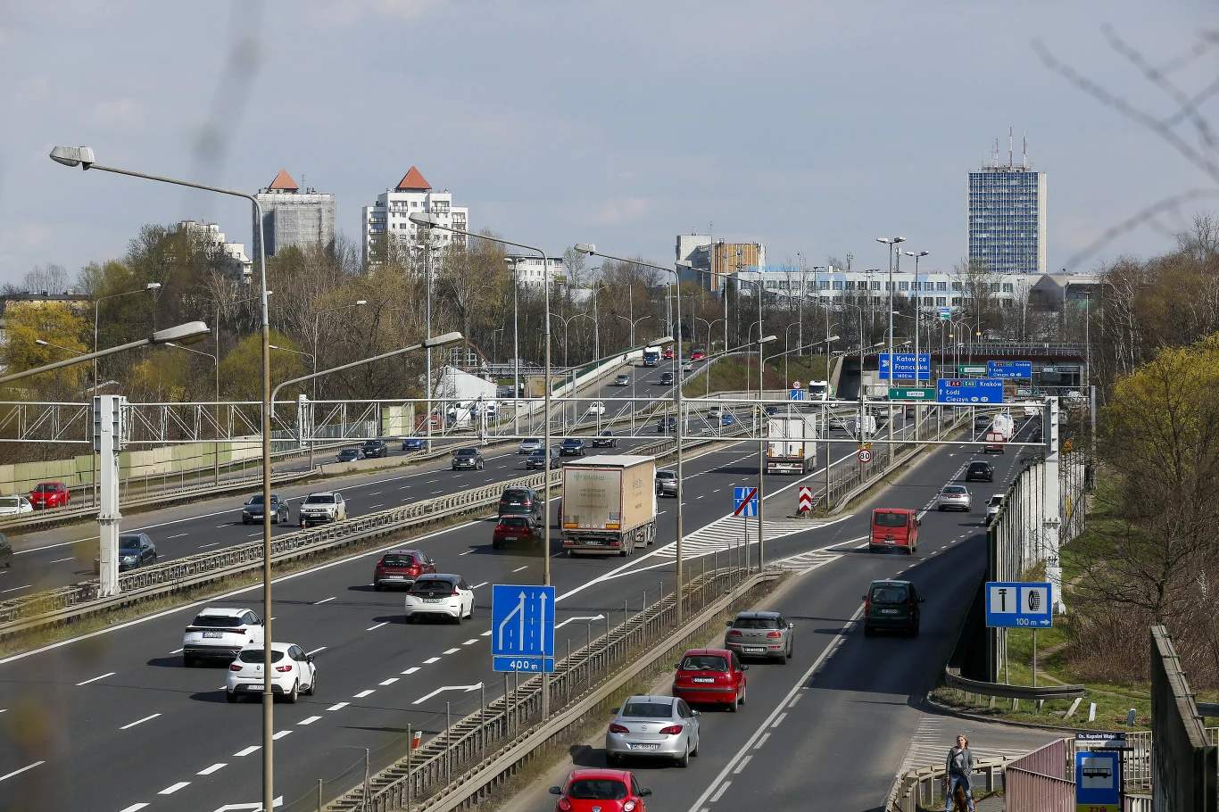 Autostrada A4 w Katowicach Ruchliwa, wielopasmowa autostrada z samochodami osobowymi i ciężarówkami, otoczona zielenią i wysokimi budynkami mieszkalnymi oraz biurowcami w tle.