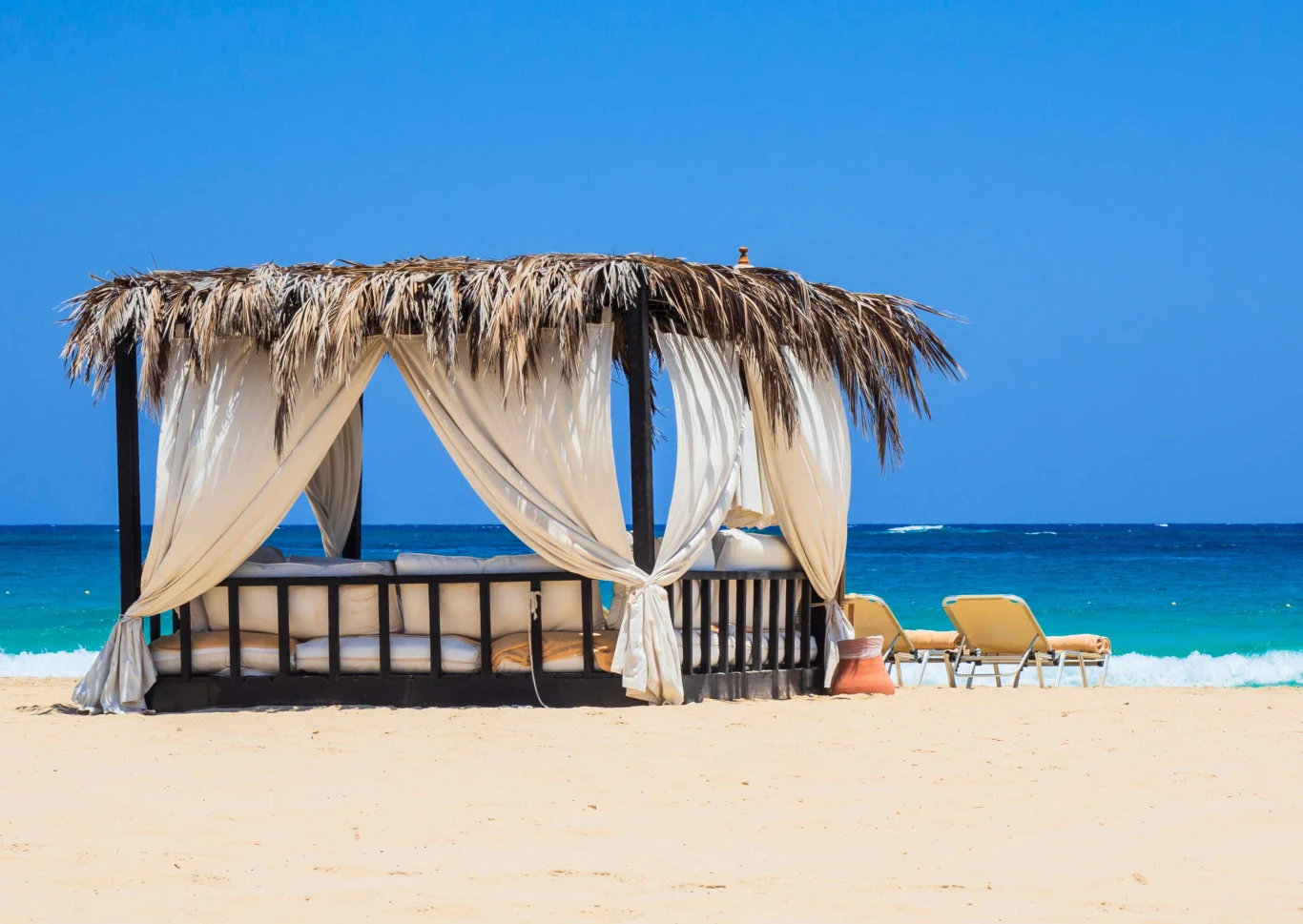 Altana z dachem z liści palmowych stoi na piaszczystej plaży tuż przy turkusowym morzu. W jej wnętrzu umieszczono wygodne łóżko, obok znajdują się dwa żółte leżaki zwrócone w stronę wody. Całość otacza błękit nieba i letnia, wakacyjna atmosfera.