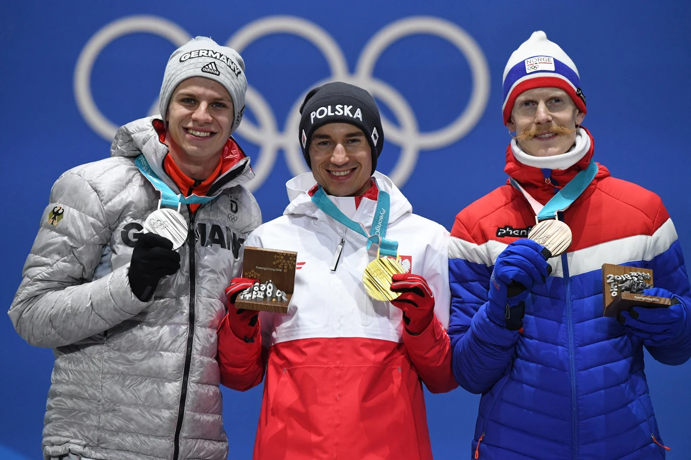 Podium zimowych igrzysk olimpijskich Pjongczang 2018: Andreas Wellinger, Kamil Stoch, Robert Johansson Trzech sportowców w kurtkach z nazwami swoich krajów, stojących na podium z medalami olimpijskimi zawieszonymi na szyjach, każdy z nich trzyma także drewniane trofeum, w tle widoczne okręgi olimpijskie.
