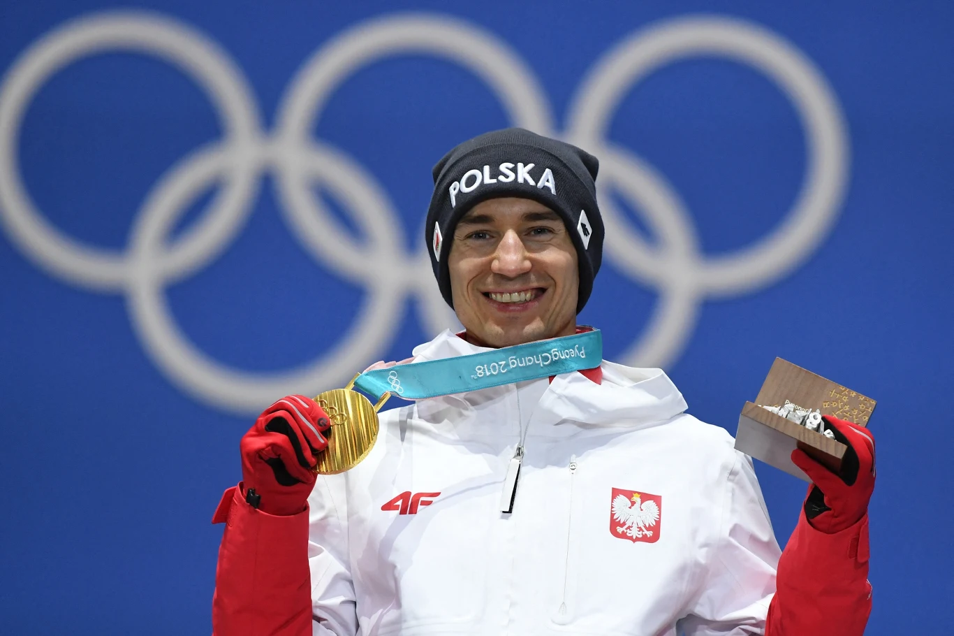 Kamil Stoch, zimowe igrzyska olimpijskie 2018 Sportowiec ubrany w biało-czerwoną kurtkę z napisem Polska, trzyma w dłoni złoty medal olimpijski i pudełko z nagrodą, w tle niebieskie tło z dużym symbolem kół olimpijskich.