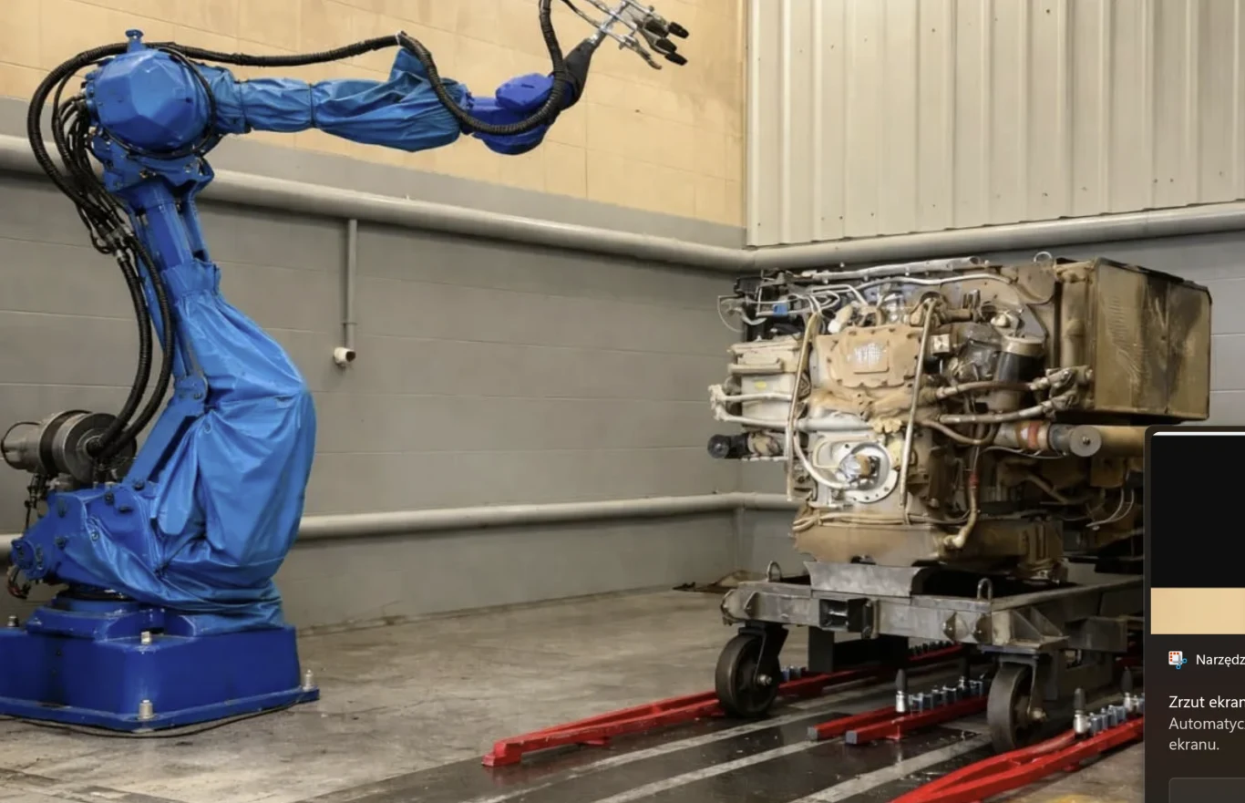 Ruby AI nie produkuje "słodkich robotów do składania prania", ale systemy rozwiązujące realne problemy armii, jak mycie i konserwacja czołgów Robot przemysłowy w niebieskim pokrowcu pracuje przy dużym silniku umieszczonym na wózku transportowym w hali produkcyjnej z szarymi ścianami.