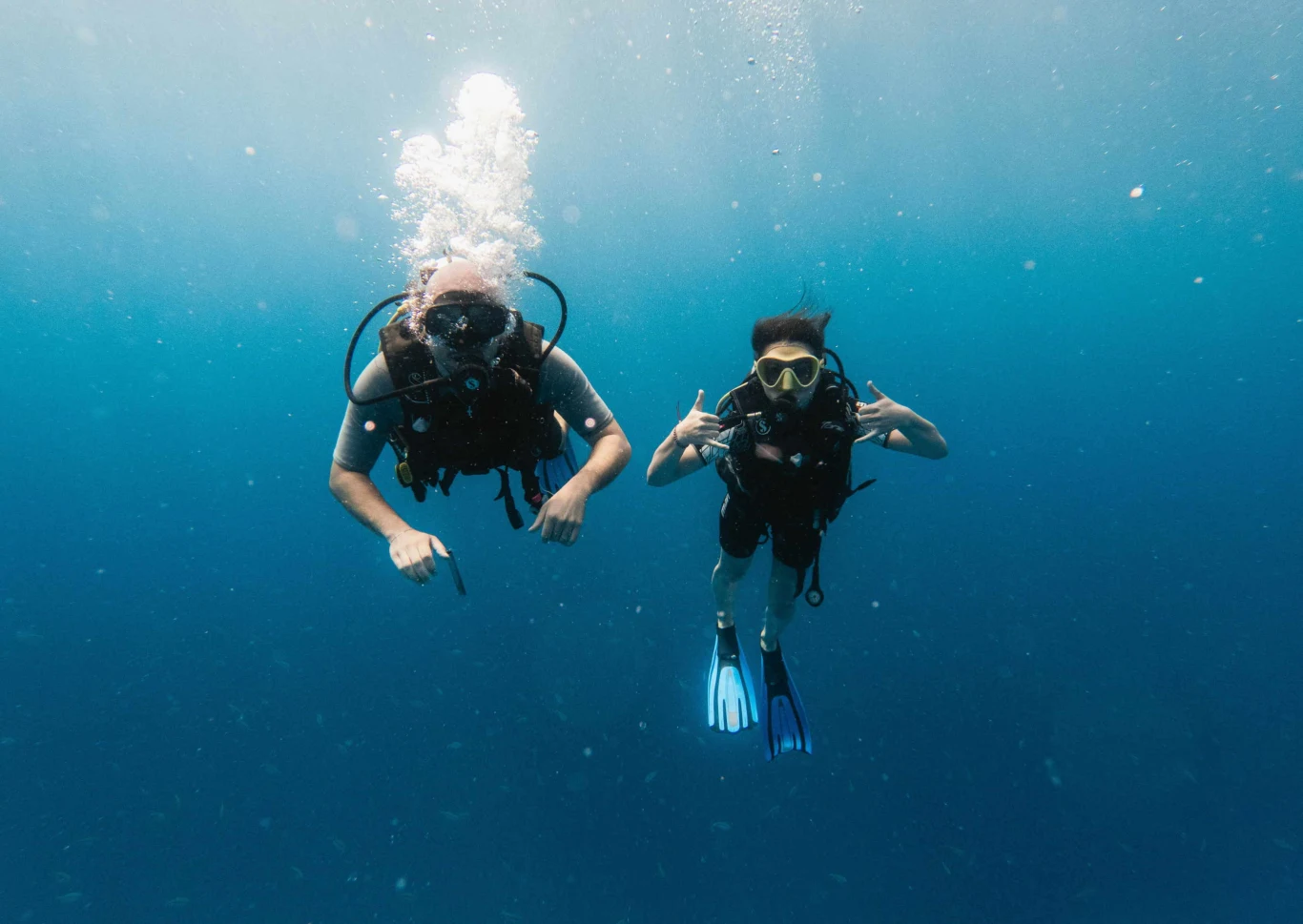 Dwoje nurków unoszących się w krystalicznie czystej, błękitnej wodzie, w pełnym ekwipunku do nurkowania, z maskami i płetwami. Jedna osoba pokazuje gesty rękami, oboje wyglądają na zrelaksowanych i cieszą się nurkowaniem.