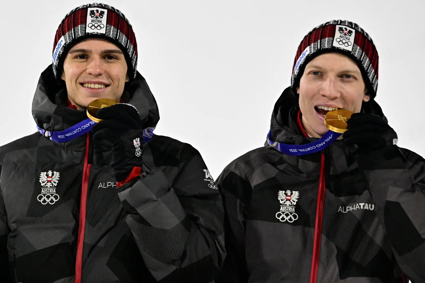 Stephan Embacher i Jan Hoerl Dwóch młodych mężczyzn ubranych w olimpijskie stroje zimowe trzyma w zębach złote medale, uśmiechając się do aparatu. Na ich kurtkach widoczne są logo reprezentacji i symbole olimpijskie, a całość utrzymana jest na jasnym tle.