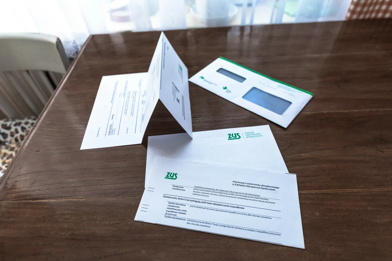 PIT-y z ZUS już trafiają do seniorów. W tych sytuacjach trzeba działać Dokumenty i koperta z logo ZUS leżące na drewnianym stole, wśród nich widoczne pismo urzędowe i otwarta koperta, całość w jasnym otoczeniu domowym.