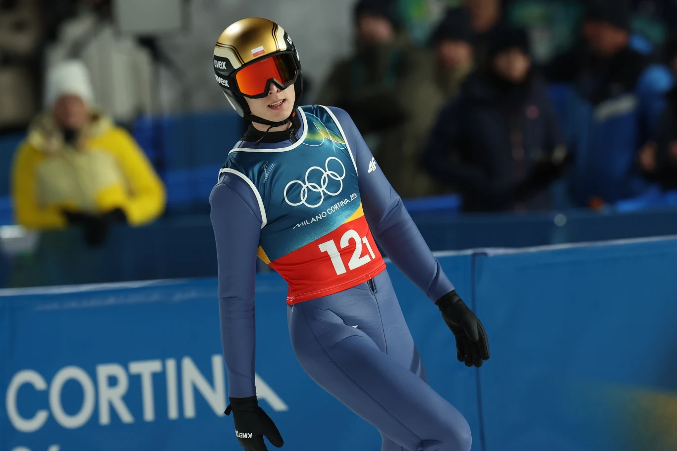 Paweł Wąsek Sportowiec w kombinezonie narciarskim i kasku ląduje po skoku podczas zawodów olimpijskich, w tle widać rozmytą publiczność i elementy infrastruktury sportowej.