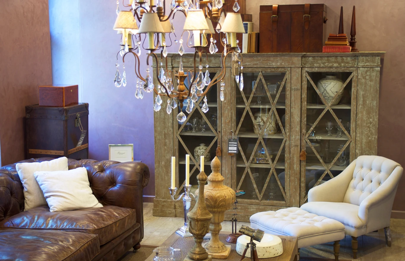 Elegancki salon w stylu vintage z brązową skórzaną sofą, białym fotelem z podnóżkiem, szklanym żyrandolem, przeszkloną witryną z dekoracjami oraz klasycznymi dodatkami i książkami.