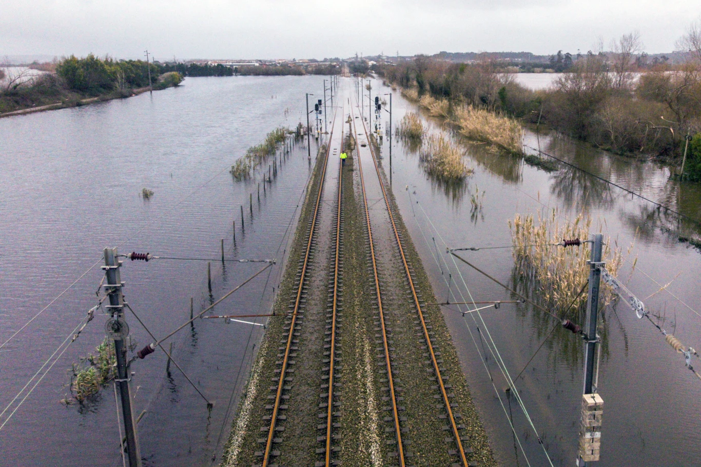 Tory kolejowe otoczone rozległym zalanym terenem, woda sięga aż do torowiska, po bokach widoczne zarośla i drzewa, pochmurne niebo potęguje atmosferę powodzi.