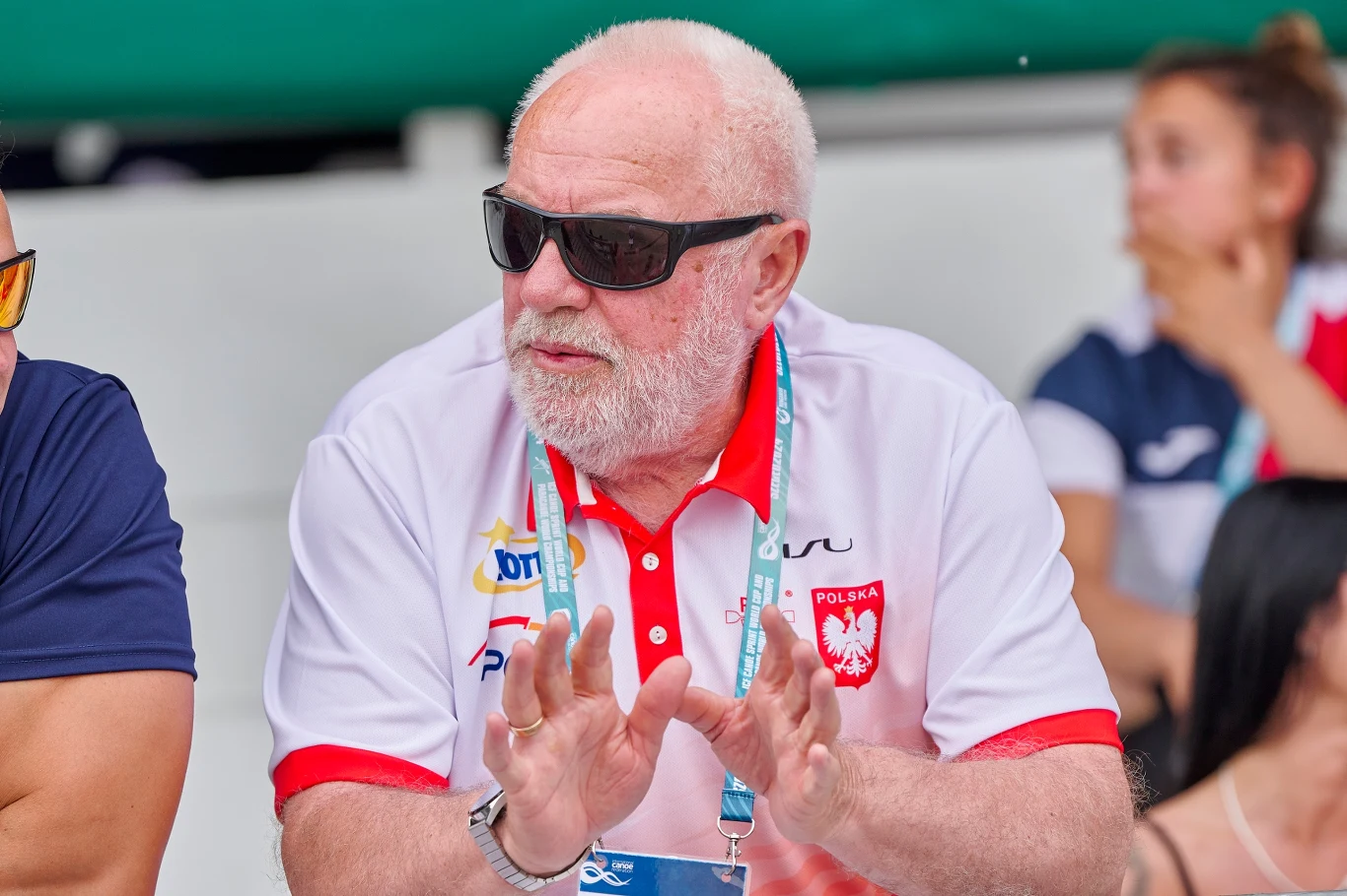 Starszy mężczyzna w okularach przeciwsłonecznych i białej koszulce polo z logo Polski, siedzi wśród innych osób podczas wydarzenia sportowego, trzymając dłonie uniesione jakby wyjaśniał coś lub kibicował.