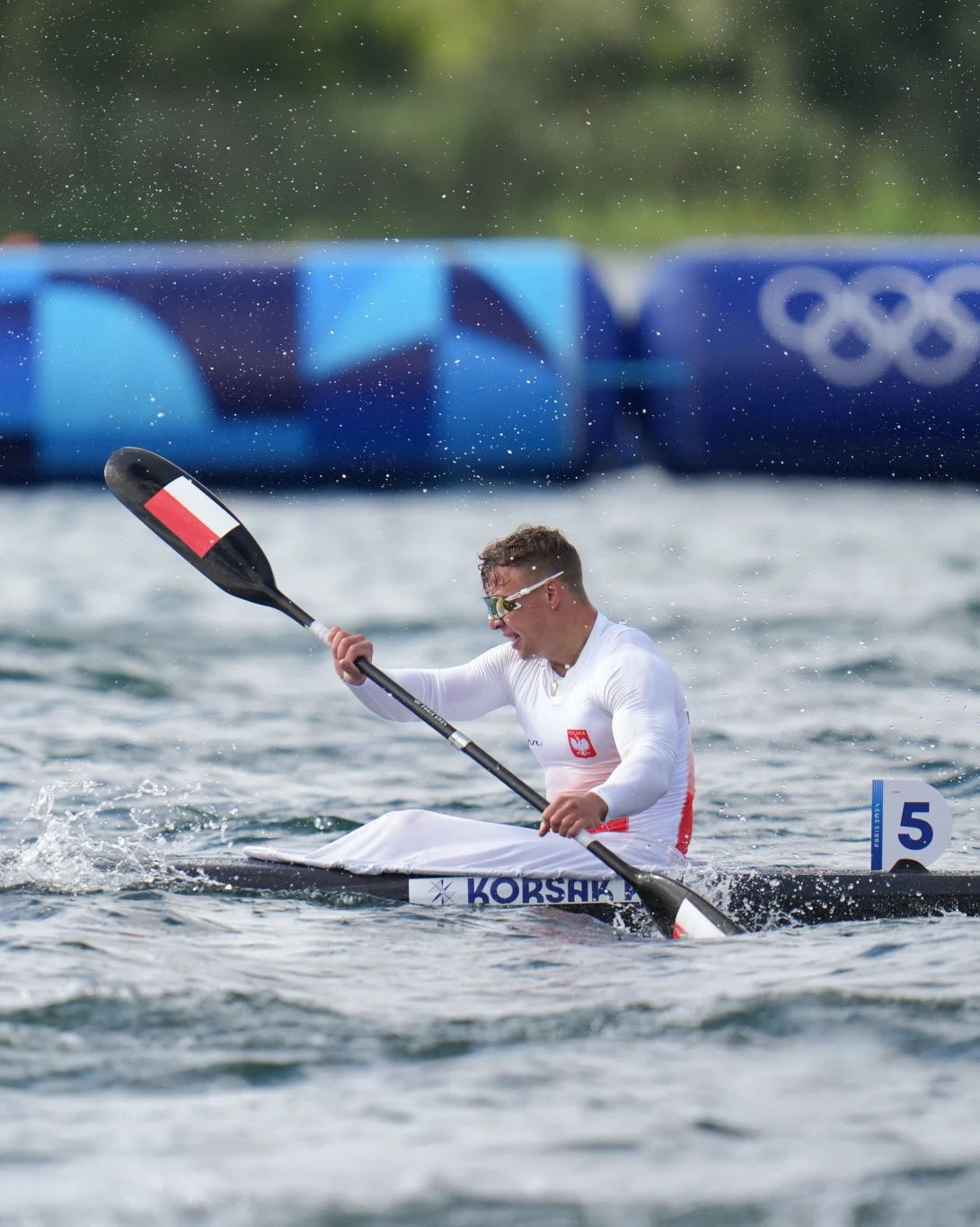 Kajakarz w białym stroju reprezentujący Polskę płynie po torze wodnym podczas zawodów olimpijskich, otoczony rozpryskami wody, z widocznym numerem 5 na łódce oraz olimpijskimi symbolami w tle.