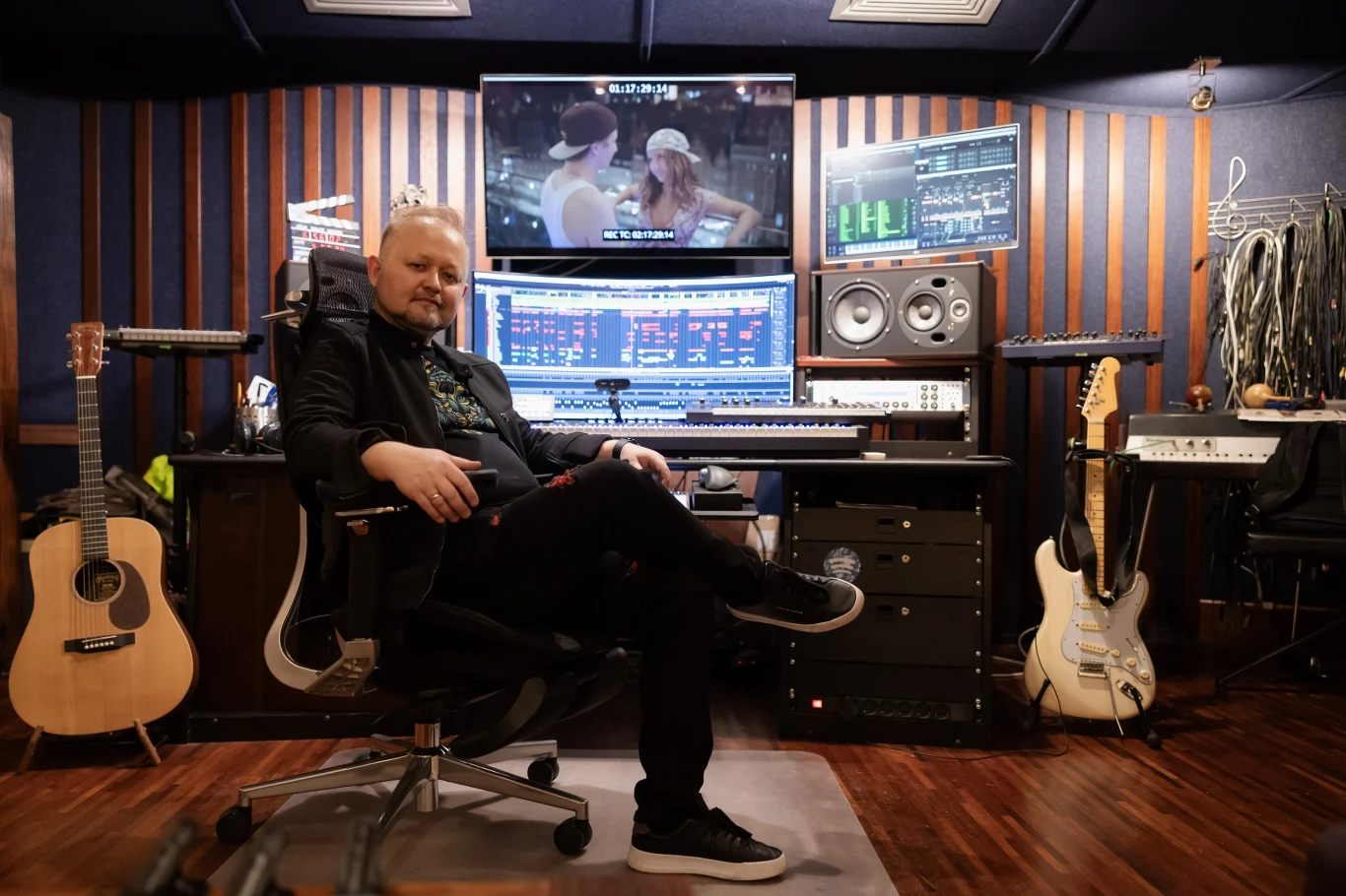 Maciej Zieliński Mężczyzna siedzący na krześle w profesjonalnym studiu muzycznym, otoczony sprzętem muzycznym, gitarami, monitorami studyjnymi oraz ekranami komputerowymi z wyświetlonym oprogramowaniem do obróbki dźwięku.