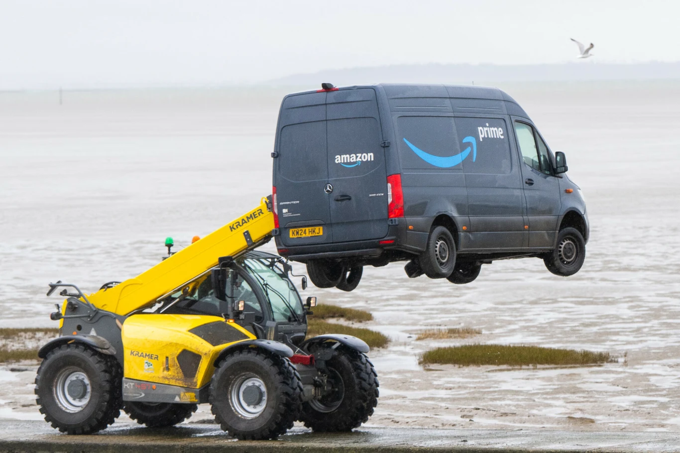 "HMS Amazon" i jego wydobycie z Tamizy. Żółty pojazd budowlany o nazwie JCB z wysięgnikiem podnosi furgonetkę dostawczą Amazon Prime nad piaskiem lub błotnistą powierzchnią, w tle pusta przestrzeń i mewa w locie.