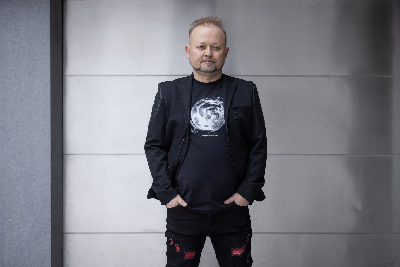 Maciej Zieliński Mężczyzna w czarnej marynarce i koszulce z nadrukiem księżyca stoi przy szarej, betonowej ścianie, z rękami wsuniętymi do kieszeni.
