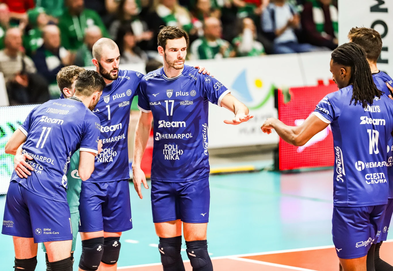 Grupa siatkarzy w niebieskich strojach drużyny zbiera się do wspólnej rozmowy na boisku podczas meczu, w tle widoczni są kibice na trybunach.