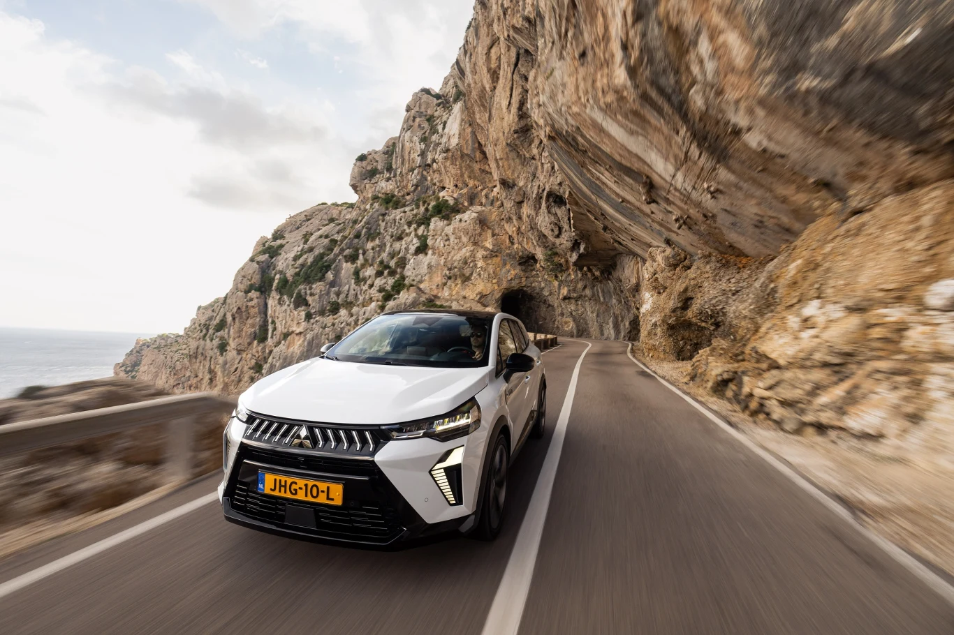 Biały SUV na zagranicznych tablicach rejestracyjnych jedzie krętą, nadmorską drogą wykutą w skalistym klifie, z widocznym tunelem oraz widokiem na morze po lewej stronie.