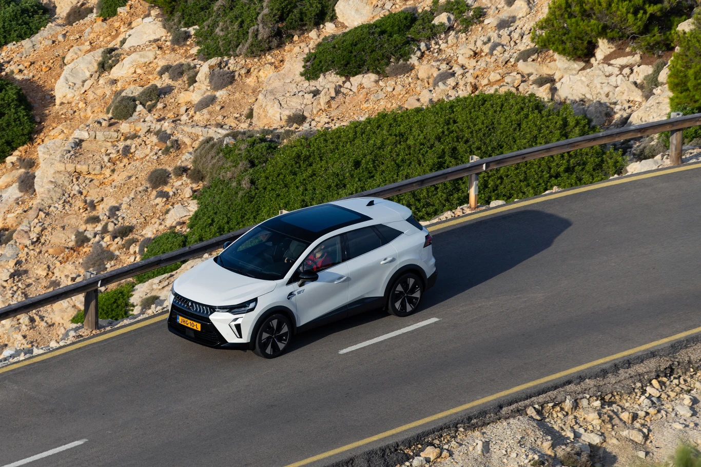 Biały samochód jedzie krętą asfaltową drogą otoczoną suchym, skalistym terenem i zielonymi krzewami, z barierką ochronną zabezpieczającą drogę