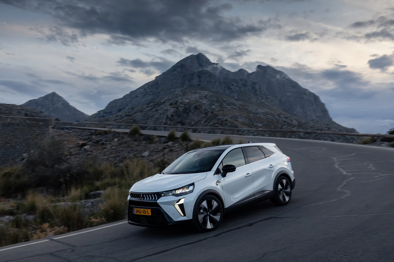 Biały nowoczesny SUV poruszający się krętą górską drogą, w tle wysokie skaliste góry i pochmurne niebo.