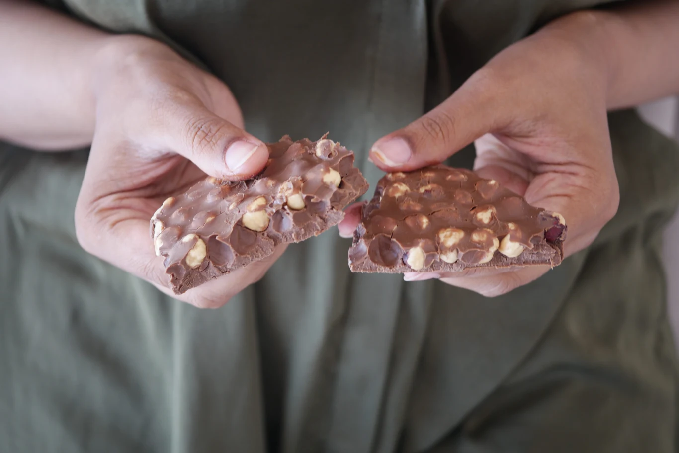 Na dobry nastrój i sen, dla serca i stabilnego cukru. Czekoladę z orzechami jedz zwłaszcza zimą Dwie ręce trzymają połamaną tabliczkę mlecznej czekolady z orzechami, widoczne kawałki czekolady oraz wyraźne orzechy na jej powierzchni, tło stanowi materiał w zielonym kolorze.