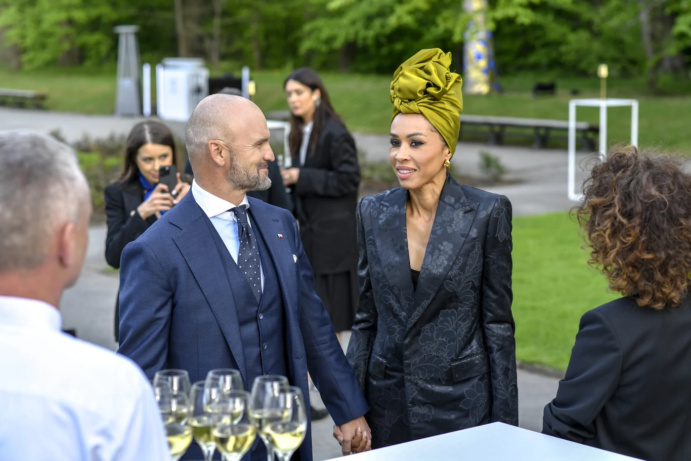 Rafał Brzoska, Omenaa Mensah Para elegancko ubranych osób trzyma się za ręce podczas towarzyskiego spotkania plenerowego, tłum w tle rozmawia ze sobą, na pierwszym planie widać kieliszki z winem ustawione na stole. Kobieta ma żółty turban na głowie i ciemną marynarkę, mężczyzna no...