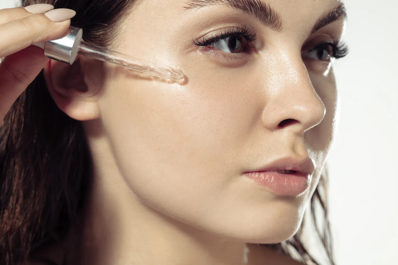 Kobieta nakłada pipetą serum na skórę twarzy, ukazując gładką cerę oraz proces pielęgnacji. Jej spojrzenie jest skupione, a światło podkreśla delikatność skóry.