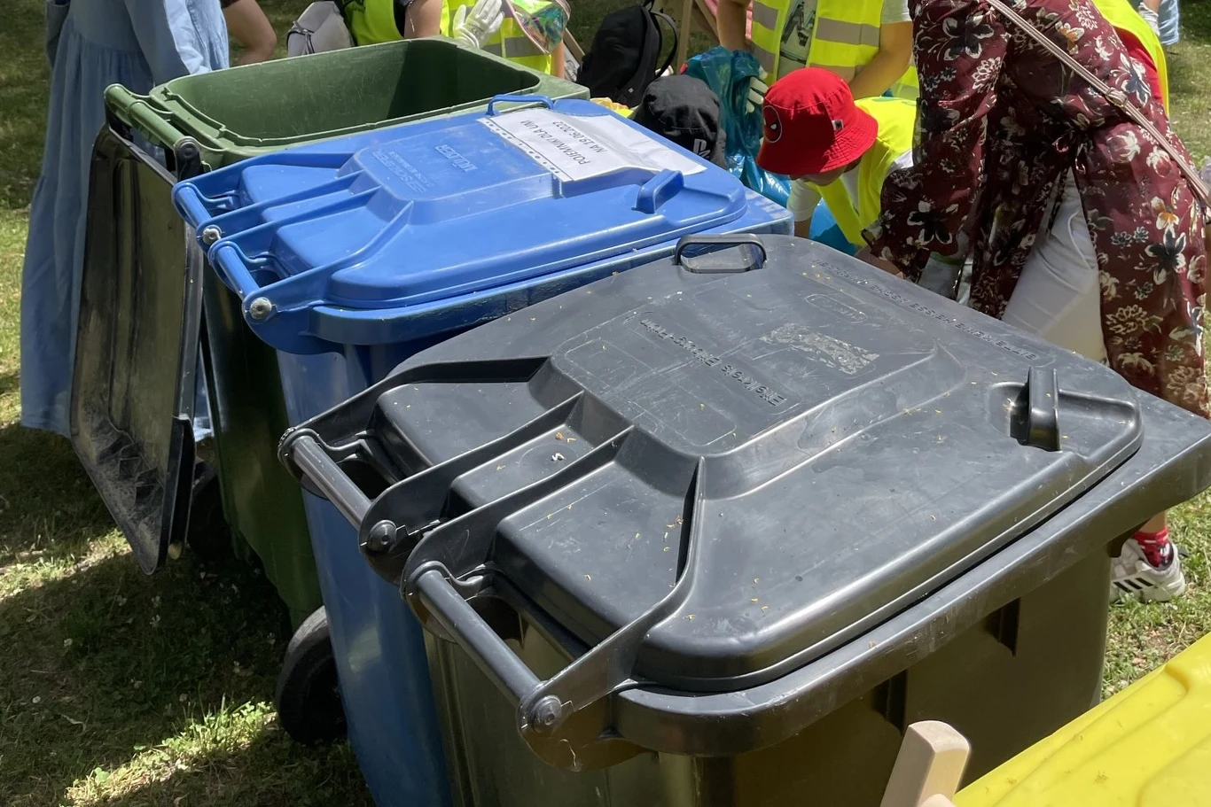 11 różnych pojemników na odpady? Kolorowe pojemniki na śmieci ustawione w rzędzie na trawie, w tle widoczne osoby w kamizelkach odblaskowych i letnich ubraniach, część z nich zajmuje się segregacją odpadów.