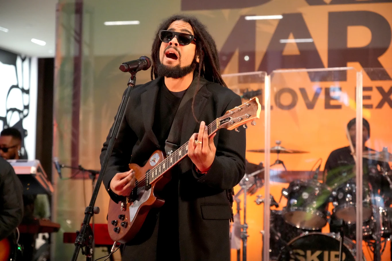 Skip Marley jest wnukiem Boba Marleya Muzyk w ciemnych okularach gra na gitarze elektrycznej i śpiewa do mikrofonu na scenie, w tle widoczny zespół i transparent z napisem.