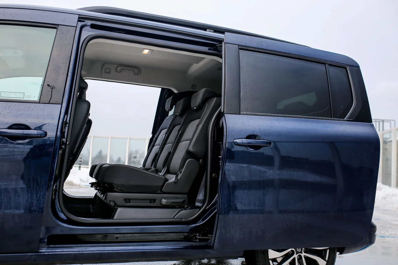 Dwie pary drzwi przesuwnych mają szerokość 83 cm (to o 18 cm więcej niż w zwykłym Kangoo). Widok wnętrza niebieskiego vana z otwartymi bocznymi drzwiami, odsłaniający trzy rzędy czarnych foteli, zaparkowanego na tle zimowego krajobrazu ze śniegiem.