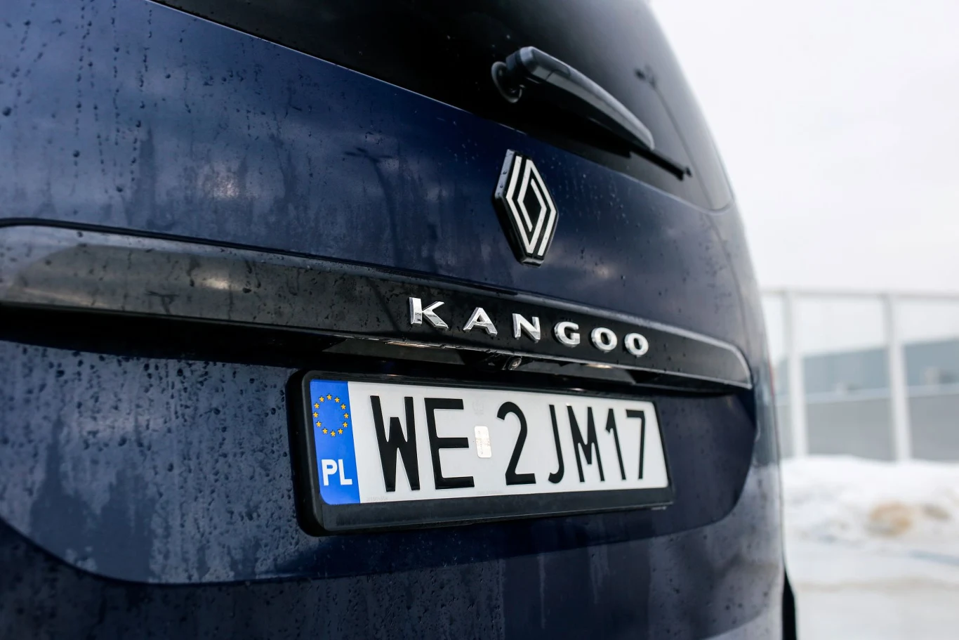 Ceny Renault Grand Kangoo zaczynają się od 132 900 zł w przypadku wariantu wysokoprężnego i od 134 900 w wersji benzynowej. Tylna część samochodu Renault Kangoo w kolorze granatowym, z widoczną tablicą rejestracyjną z Polski i emblematem marki na klapie bagażnika. Na powierzchni pojazdu są ślady deszczu, w tle widoczny fragment śniegu.