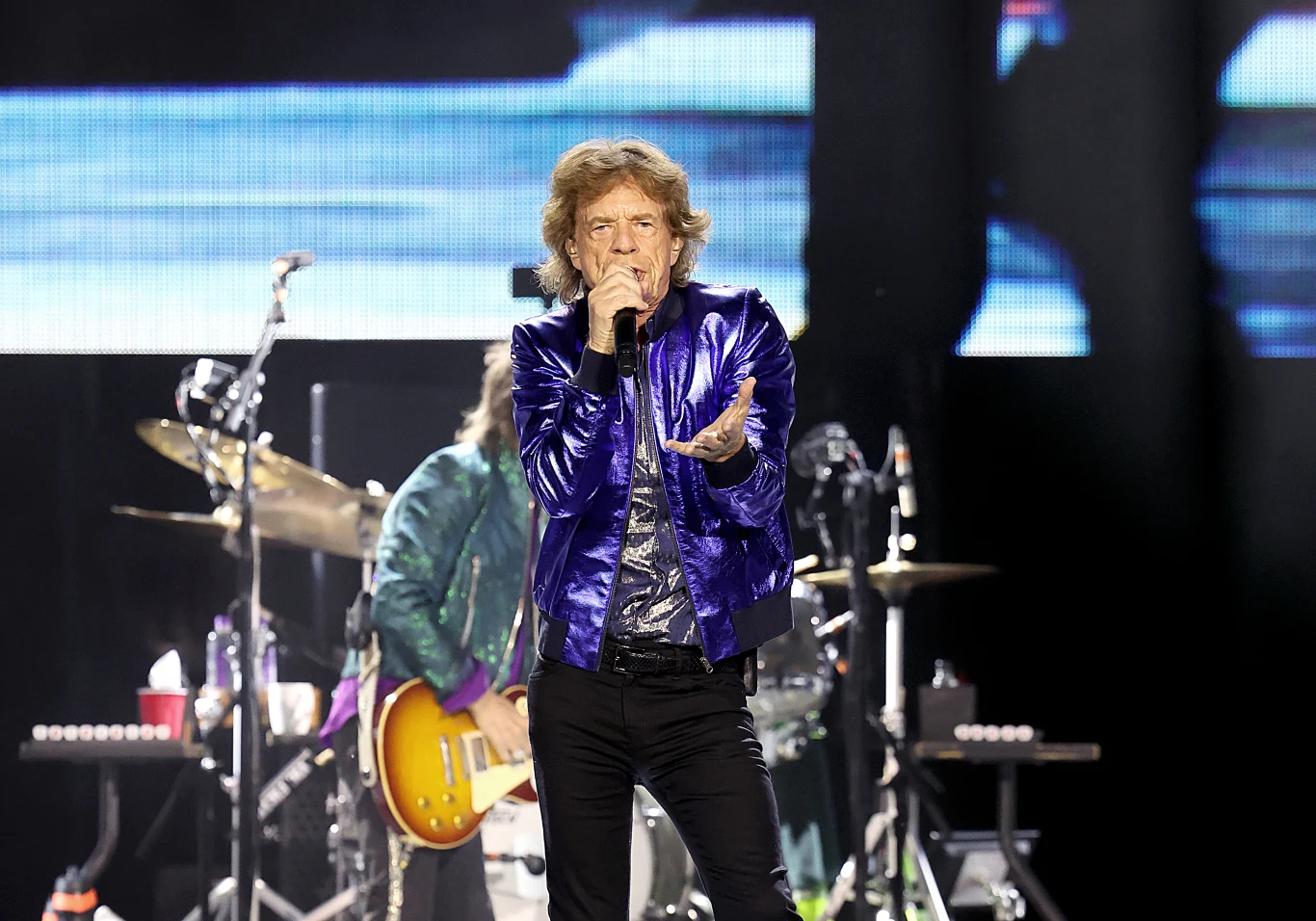 Mick Jagger, wokalista The Rolling Stones Wokalista w niebieskiej, błyszczącej kurtce śpiewający na scenie koncertowej, w tle widoczny gitarzysta z elektryczną gitarą i zestaw perkusyjny, dynamiczne oświetlenie sceniczne.